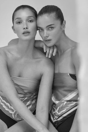 Close-up portrait of two female models with sleek pulled-back hair posing together against a plain studio background.