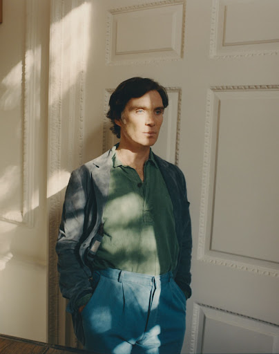 Portrait of fair-skinned adult male with dark hair in a green shirt and blue pants, standing indoors near a paneled door with sidelight.