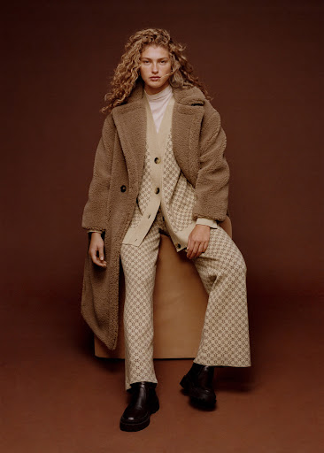 Adult woman with curly hair sits in a patterned suit and teddy coat, posing in front of a brown background in a studio setting.