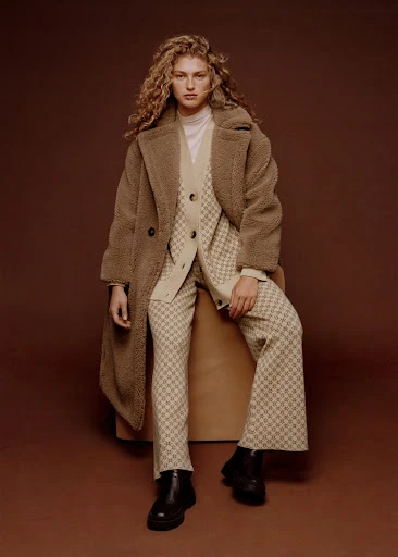 Adult woman with curly hair sits in a patterned suit and teddy coat, posing in front of a brown background in a studio setting.
