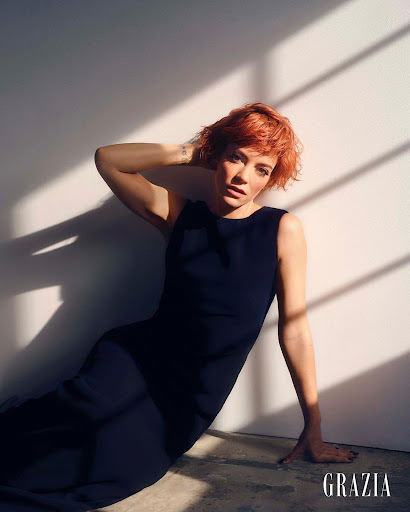 Elegant woman with short red hair in a sleeveless dress leans against a wall with sunlight and shadows in a fashion editorial shot.