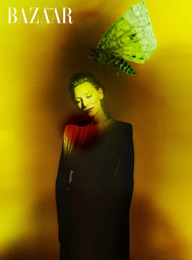 A woman in a black outfit poses with a large moth against a textured yellow background in a fashion editorial image. 