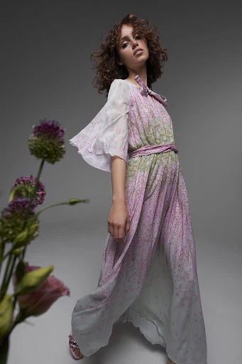 A young woman with curly hair poses in a flowing dress with floral accents in a studio environment with diffused lighting.