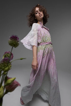 A young woman with curly hair poses in a flowing dress with floral accents in a studio environment with diffused lighting.