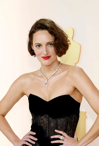 Close-up of Phoebe Waller-Bridge at an event, wearing a black strapless dress, a statement necklace, and red lipstick, hands on hips.