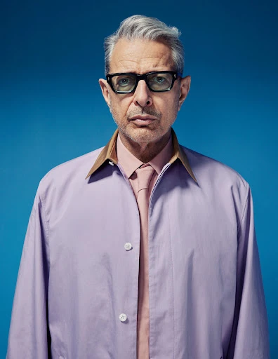 Jeff Goldblum portrait in light lavender jacket, pink shirt and tie against a blue backdrop, wearing square black glasses, medium shot