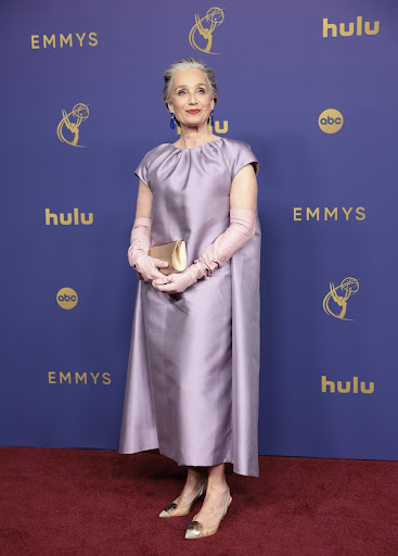 Older woman in a lavender dress and opera gloves poses on a red carpet in front of a blue backdrop with logos at an awards show.