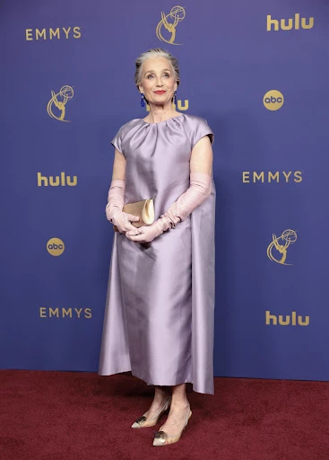 Older woman in a lavender dress and opera gloves poses on a red carpet in front of a blue backdrop with logos at an awards show.