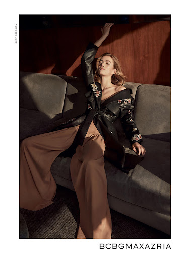 Fashion shot of a blonde adult female model lounging on a grey sofa wearing tan pants and a black floral-embroidered jacket indoors.