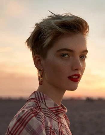 Close-up portrait of a woman with short blonde hair, red lipstick, and plaid shirt against a sunset sky.