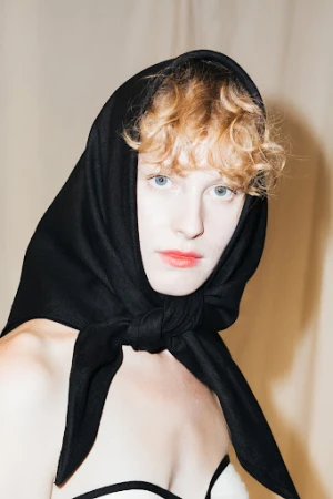 Portrait of a woman with curly blonde hair and blue eyes wearing a black headscarf tied under her chin.