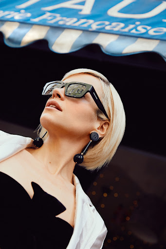 Blonde woman wears oversized sunglasses and black dress with statement earrings under an awning, fashion editorial lifestyle shot.