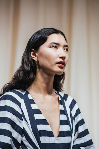 Close-up portrait of a woman with dark wavy hair and red lipstick wearing a blue and white striped V-neck top against fabric backdrop.