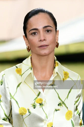 Woman in a dandelion-print shirt, looking at the camera with a neutral expression, wearing heart-shaped earrings. Head and shoulders shot.