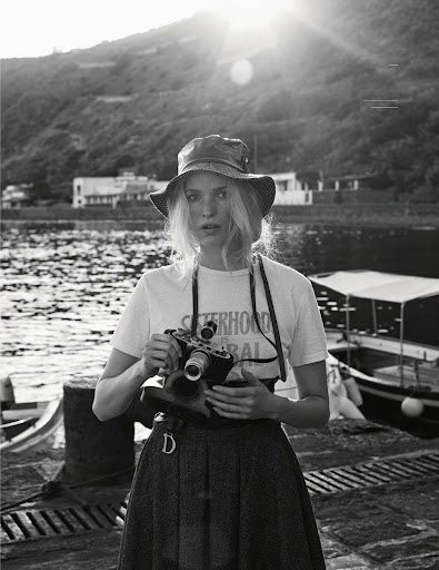 Black and white portrait of a fair-skinned woman in a hat holding a camera, with a mountain and water in the background.