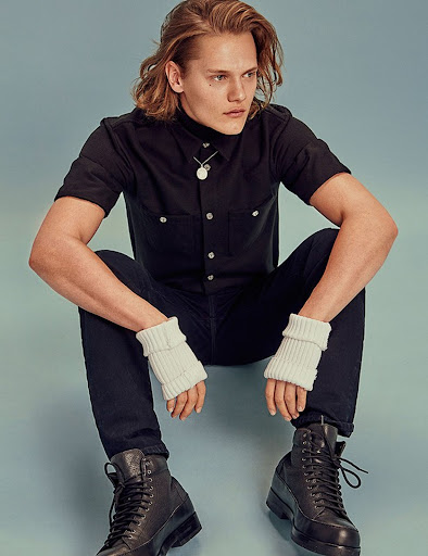 Man with long hair poses in a black button-down shirt, dark pants, and lace-up boots, sitting in a squat position on a light blue background.