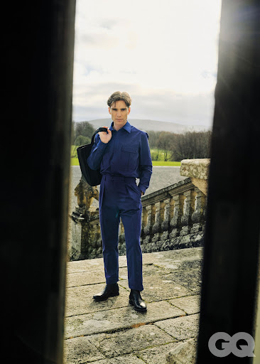 Cillian Murphy wearing a blue jumpsuit and holding a dark bag while standing on stone steps outdoors.