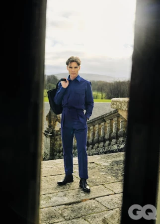 Cillian Murphy wearing a blue jumpsuit and holding a dark bag while standing on stone steps outdoors.