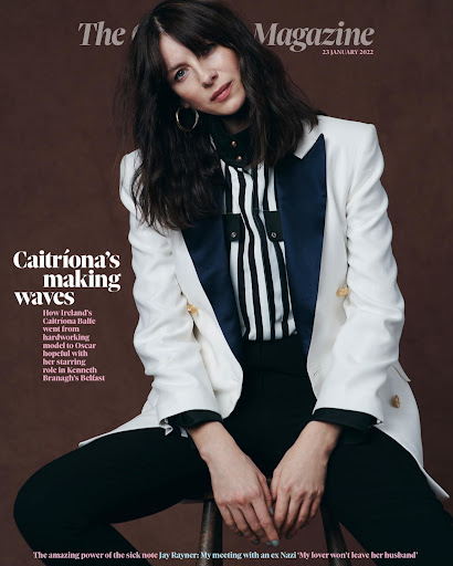Caitríona Balfe magazine cover. Woman seated, wearing a white blazer, striped shirt, and black pants, set against a brown backdrop.