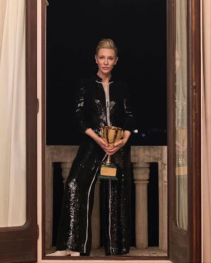 Cate Blanchett stands in a doorway, wearing a black sequin outfit and holding a gold trophy, against a dark outdoor background.