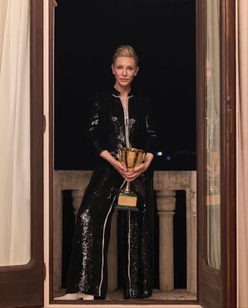 Cate Blanchett stands in a doorway, wearing a black sequin outfit and holding a gold trophy, against a dark outdoor background.