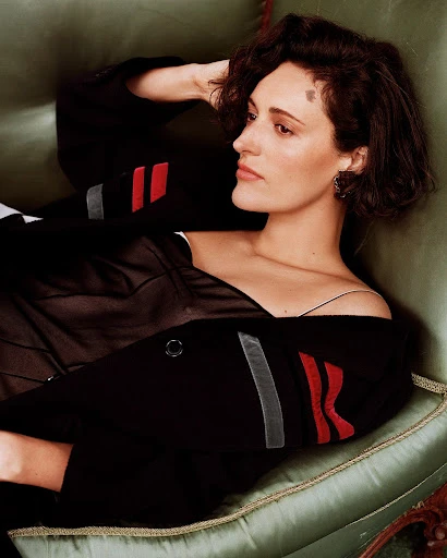 Phoebe Waller-Bridge lies on a vintage couch wearing a black jacket with red and grey stripes, staring intensely into the camera.