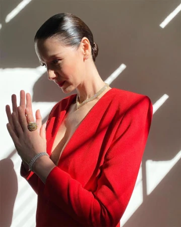 Woman in red v-neck dress with slicked-back hair wearing a diamond necklace and ring, posing against patterned light and shadow.