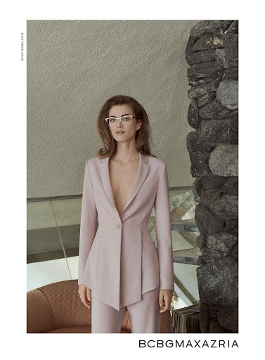 Fashion editorial shot of a woman in a light pink suit and glasses, posing indoors near a stone accent wall. BCBGMAXAZRIA brand displayed.