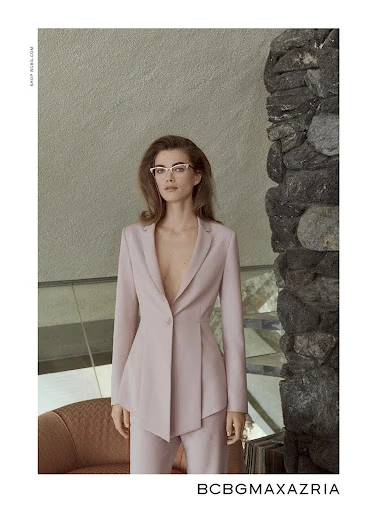Fashion editorial shot of a woman in a light pink suit and glasses, posing indoors near a stone accent wall. BCBGMAXAZRIA brand displayed.