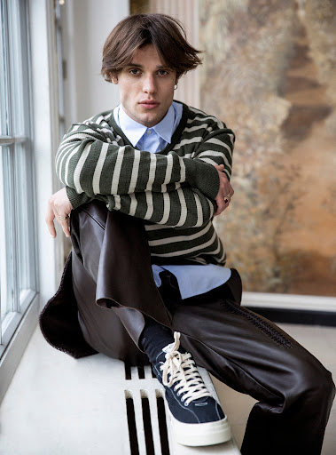Young man in striped sweater poses seated by window in relaxed fashion shot, showcasing a casual yet stylish look indoors.
