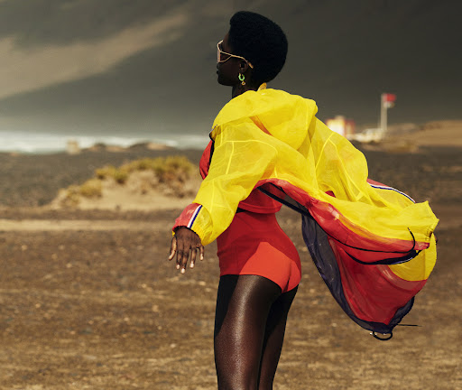 Woman with dark skin and an afro poses in a red swimsuit and yellow windbreaker jacket against a beach and stormy sky backdrop.