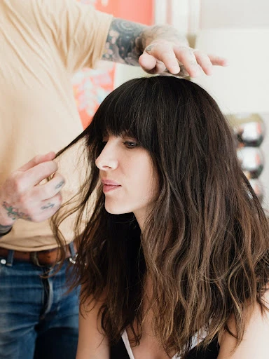 Brunette woman with wavy hair is being styled; a hairstylist with tattoos is shown. The indoor scene features fair skin and a side view.