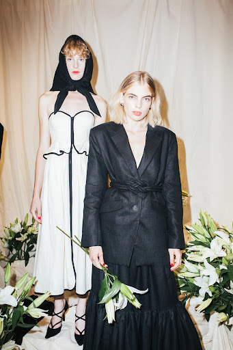 Two women pose in a studio wearing a blazer, dress and headscarf. White flowers in the foreground complement the cream-colored background.