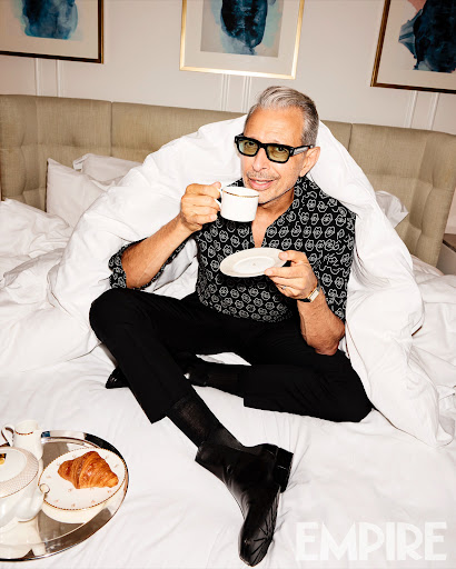 Jeff Goldblum poses in a bed with a tea set, wearing a patterned shirt and glasses, indoors with artwork in the background.
