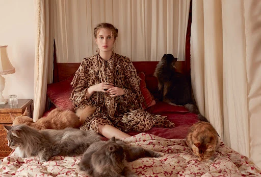 A woman in a leopard print dress sits on a bed surrounded by several cats, creating a surreal and intimate portrait scene.