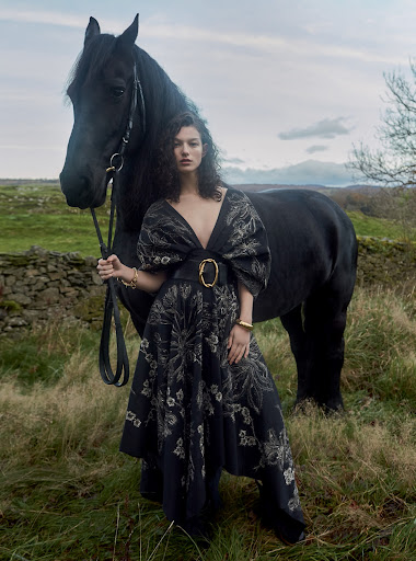 Elegant woman in a long floral dress poses with a black horse in a rural landscape, showcasing fashion, nature, and classic style.