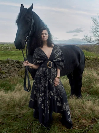 Elegant woman in a long floral dress poses with a black horse in a rural landscape, showcasing fashion, nature, and classic style.