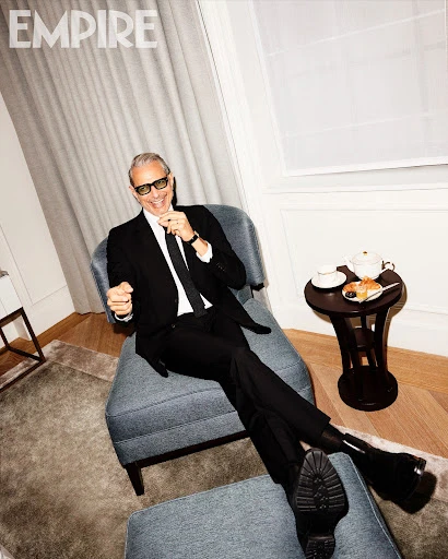 Mature adult man in a dark suit, wearing glasses, seated in blue chair with a side table and tea set indoors, magazine cover look.