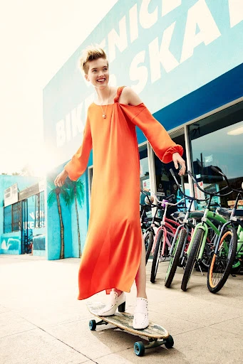 A young woman with short hair, wearing an orange dress, smiles while skateboarding in front of a bike rental shop on a sunny day.