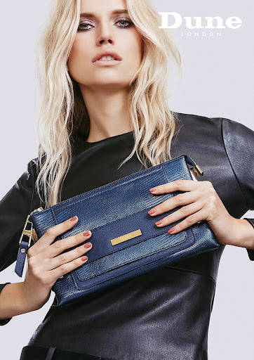 Fashion shot of a blonde model holding a blue Dune London handbag against a neutral background, emphasizing the bag's style and texture.
