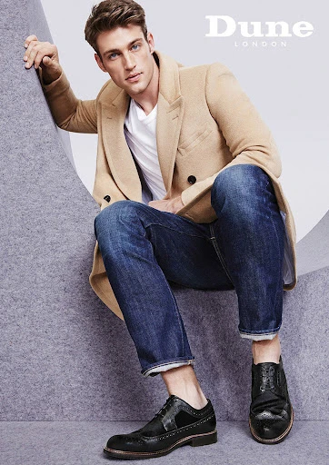 A male model poses while sitting in a beige coat, blue jeans, and black dress shoes, alongside Dune London brand text.
