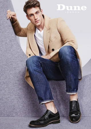 Man wearing camel coat, white t-shirt, dark jeans, and black shoes seated against grey structural set backdrop