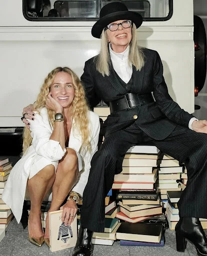 Two women sitting on a stack of books. One in a pinstripe suit and hat, the other in a white jacket and gold heels, smiling.