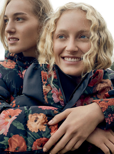 Two fair-skinned women with blonde hair wear matching floral-print puffer jackets, one smiling at the camera in a medium close-up.