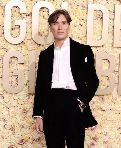 Cillian Murphy poses on the red carpet in a dark tuxedo and white shirt. He stands in front of a floral backdrop at an event.