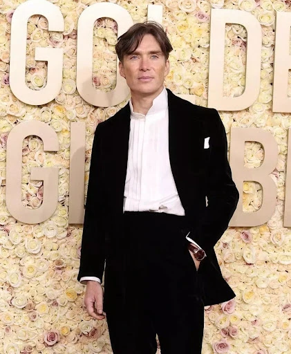 Cillian Murphy poses on the red carpet in a dark tuxedo and white shirt. He stands in front of a floral backdrop at an event.