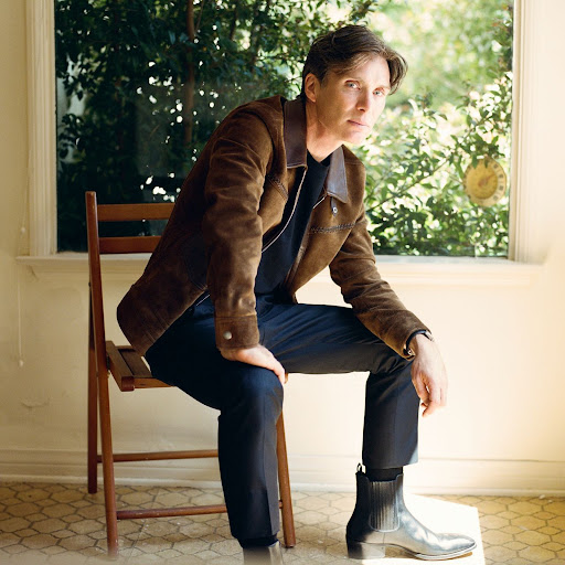 Portrait of a fair-skinned adult male with dark hair, wearing a brown jacket, sitting on a wooden chair indoors near a window.