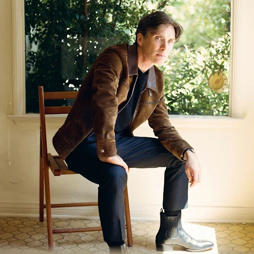 Portrait of a fair-skinned adult male with dark hair, wearing a brown jacket, sitting on a wooden chair indoors near a window.