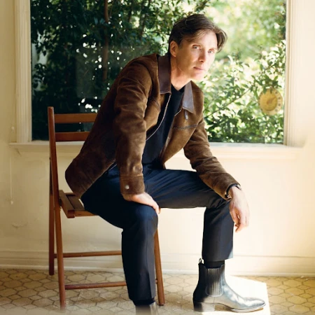 Portrait of a fair-skinned adult male with dark hair, wearing a brown jacket, sitting on a wooden chair indoors near a window.