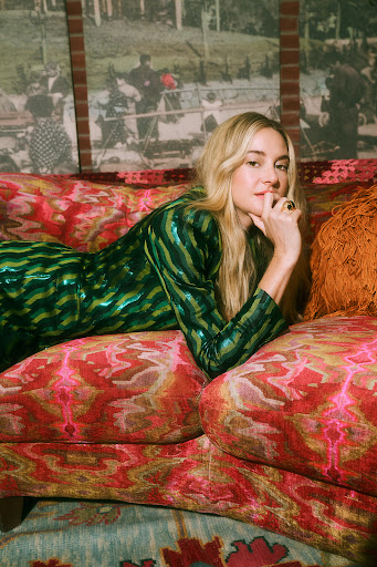 Adult female with blonde hair lies on a patterned sofa indoors, wearing a shiny green patterned dress, hand on chin, looking at the camera.